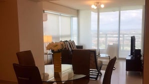 Un salon moderne avec une table à manger en verre, des sièges confortables, un téléviseur et une grande fenêtre donnant sur la ville.