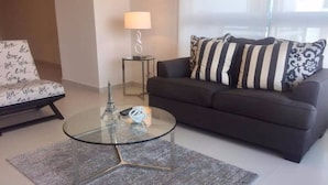 Un salon moderne avec une table basse en verre, un canapé foncé agrémenté de coussins à rayures, une petite table d’appoint avec une lampe et un fauteuil avec un coussin à motifs.