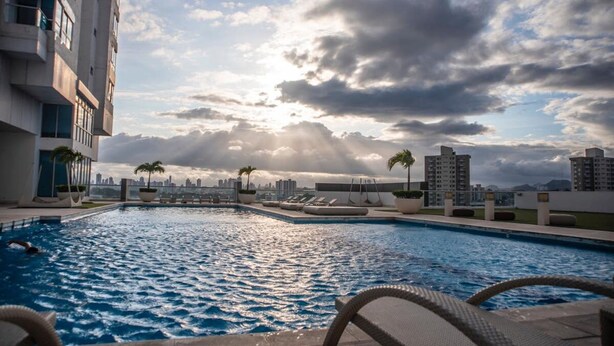 Una piscina en la azotea con sillones de descanso y palmeras, rodeada de rascacielos y un cielo nublado.