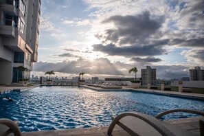 Una piscina en la azotea con sillones de descanso y palmeras, rodeada de rascacielos y un cielo nublado.