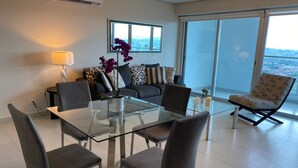 Una sala de estar moderna con una mesa de comedor de vidrio, sillas tapizadas en gris, un sofá con cojines estampados y un amplio ventanal con vistas a la ciudad.