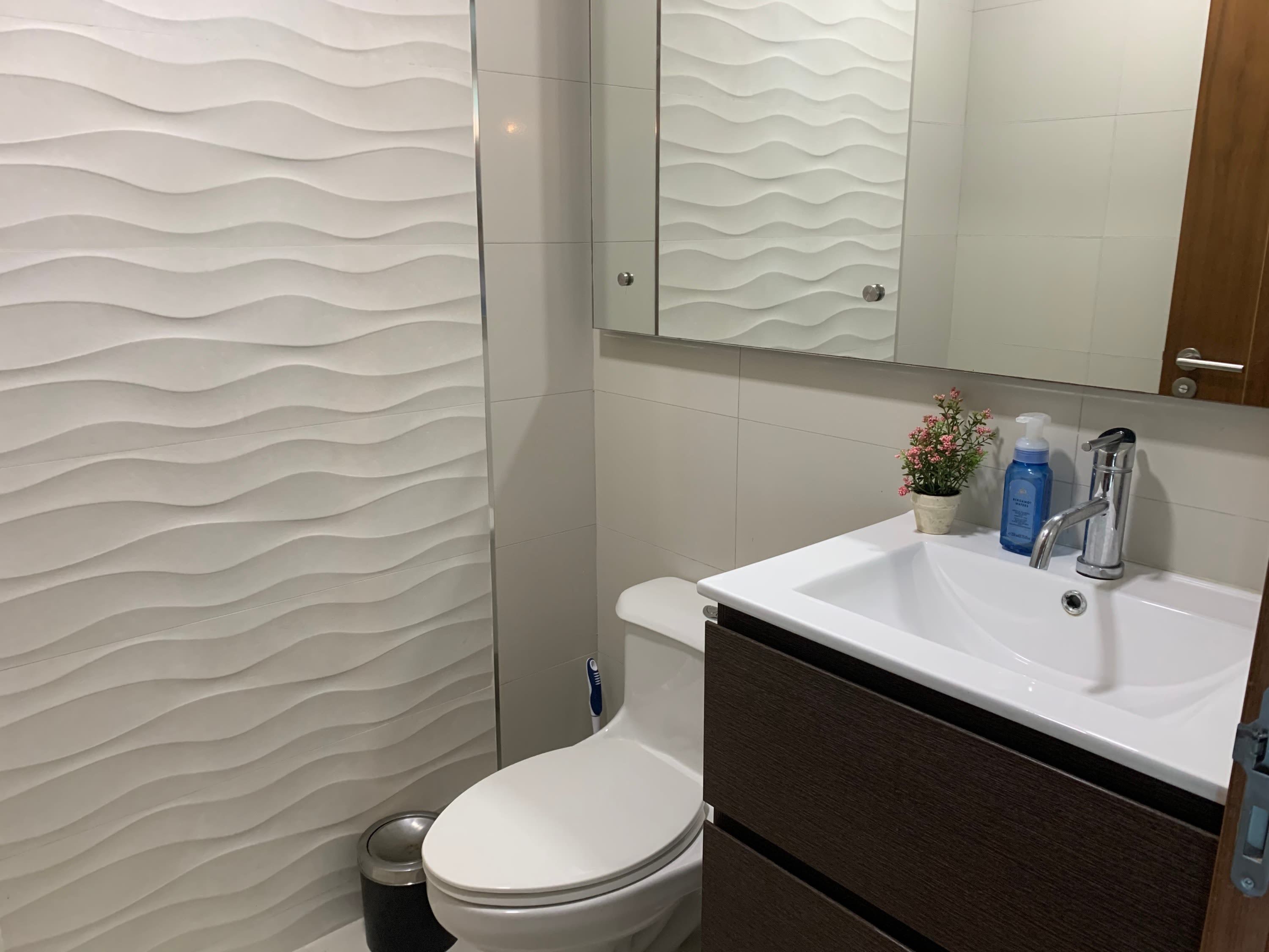 Ein modernes Badezimmer mit weißer Wand mit Struktur, einer weißen Toilette, einem Waschbecken mit Spiegel darüber und einem kleinen Topfpflanz auf dem Waschbecken.