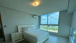 A modern bedroom with a large bed, a bedside table, a nightstand, a window with a city view, and an air conditioning unit. - Pacific Cafe Classy 1-bedroom in Costa del Este (Panama City)