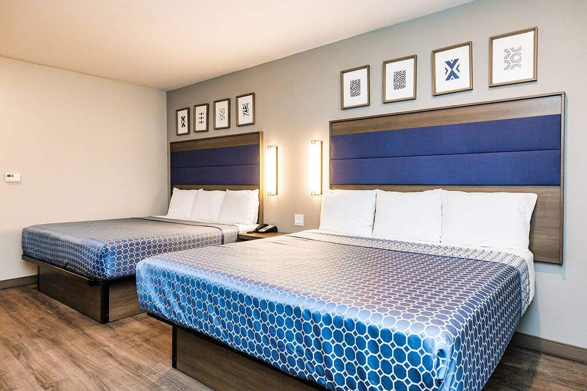 Habitación de hotel con dos camas, cada una con cabecero, mesita de noche y lámpara. Hay cuadros enmarcados en la pared.