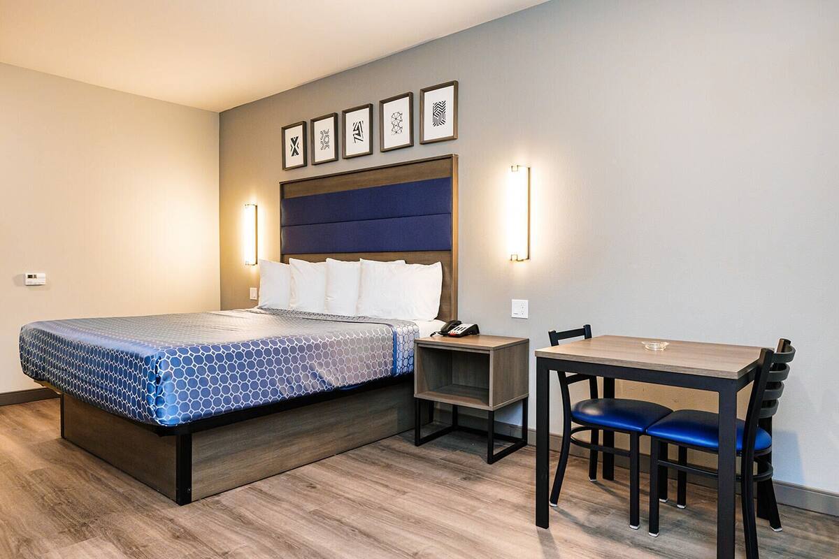 Habitación de hotel con cama, mesita de noche, silla y escritorio con otra silla. Hay luces fijadas a la pared y cuadros enmarcados.