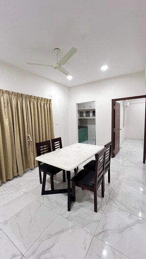 A dining area with a white table and chairs, a ceiling fan, and a kitchen in the background. - LUSHVISTAS Serviced Apartment (Hyderabad)