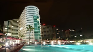 A hotel with a curved, multi-story structure, a pool area with sun loungers, and a palm tree.