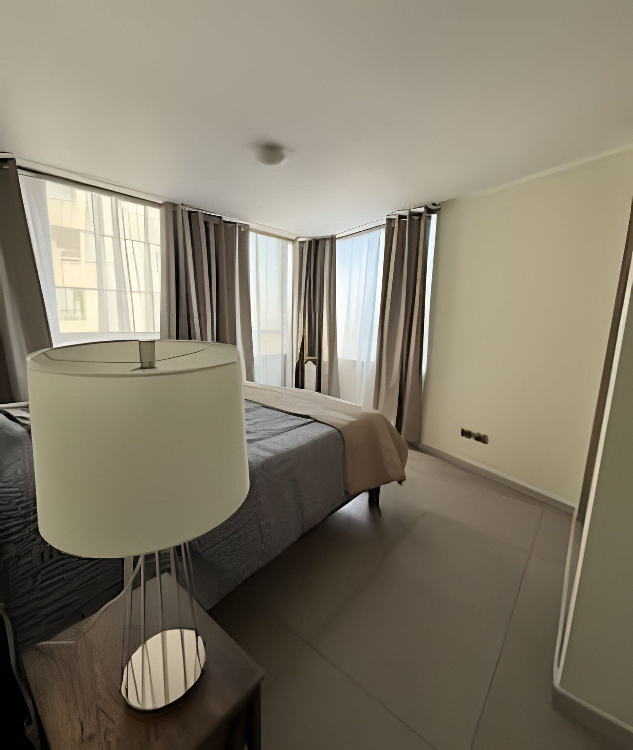 Ein Hotelzimmer mit Bett, Nachttisch mit Lampe, Fenster mit Vorhängen und Blick ins Freie.