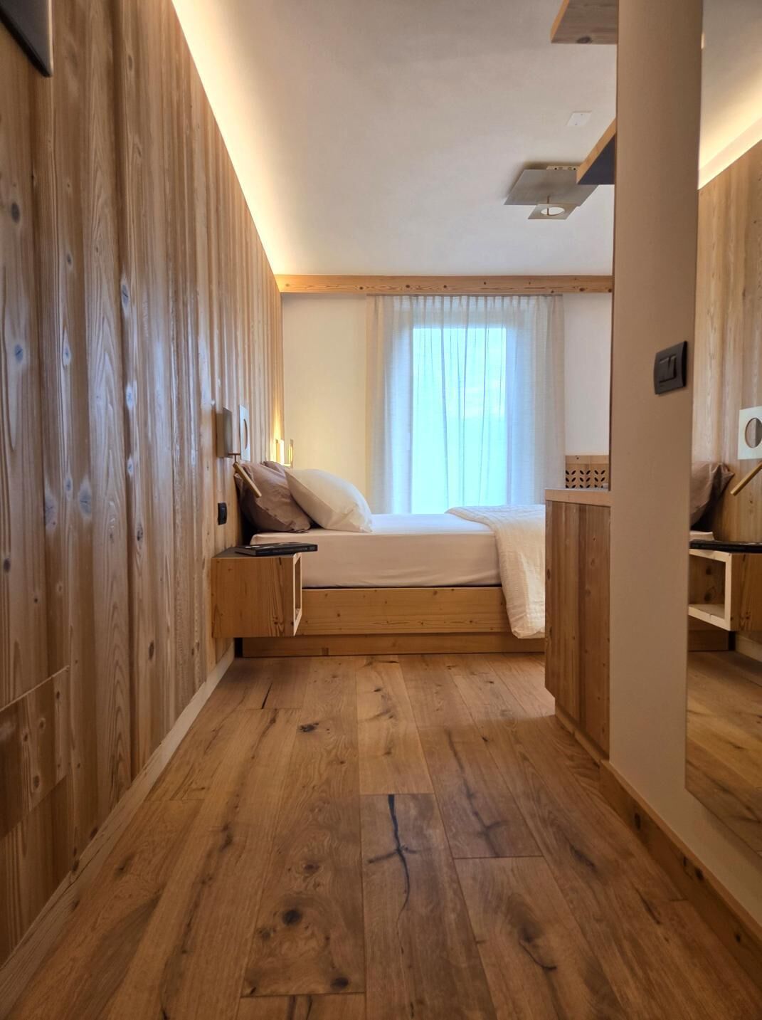 Un dormitorio con piso de madera, una cama con colchón y almohadas blancas, una mesita de noche y una ventana con cortinas.