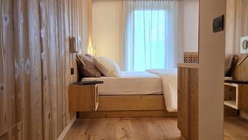 Un dormitorio con piso de madera, una cama con colchón y almohadas blancas, una mesita de noche y una ventana con cortinas.