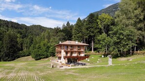 A two-story chalet with a balcony, surrounded by a grassy field and forested hills. - Il Soffio - Cucina & Camere nel bosco (San Lorenzo Dorsino)
