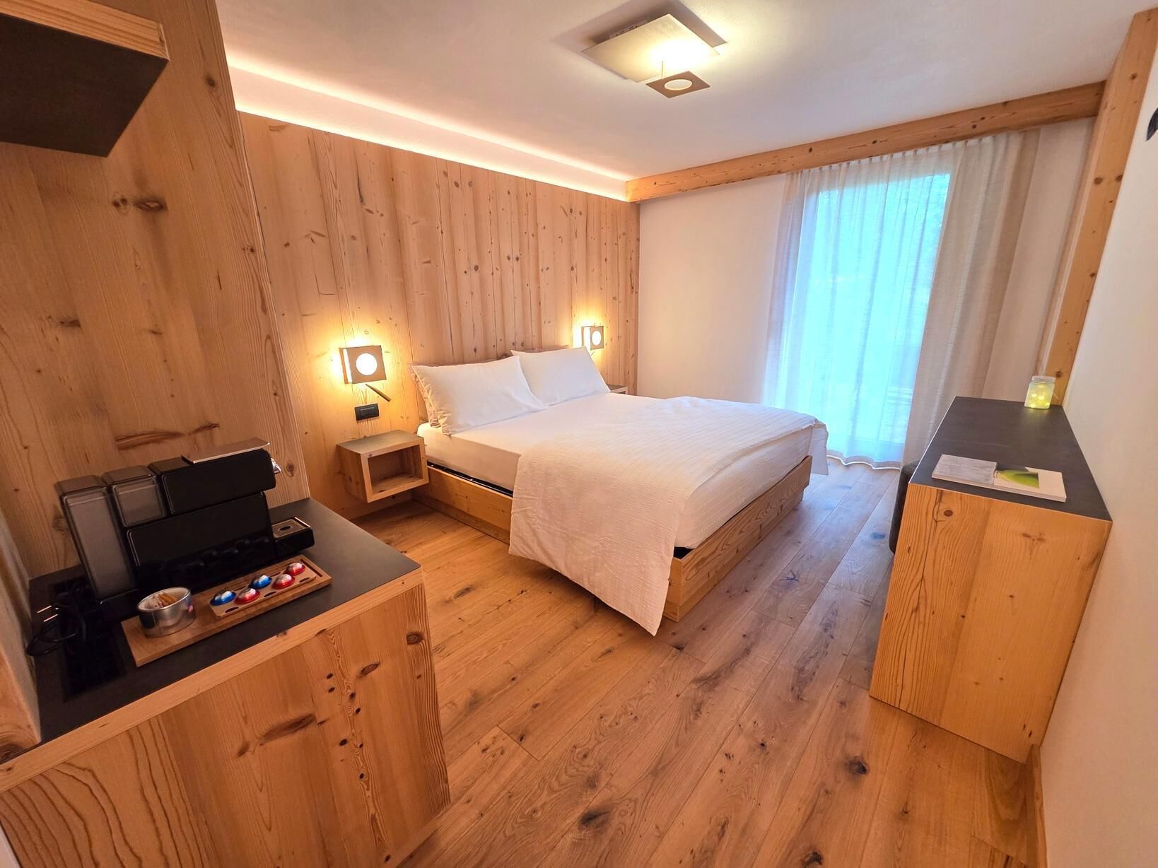 Una habitación con paneles de madera, una cama, mesitas de noche, un escritorio con una computadora y un ventanal con cortinas.