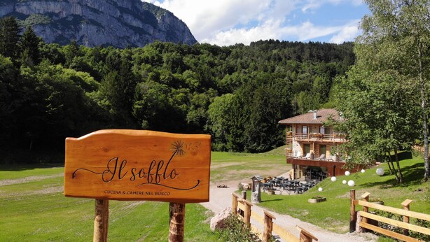 A wooden sign with "Il Solco" and "Cucina & Camera nel Bosco" text, a mountain backdrop, and a building with outdoor seating. - Il Soffio - Cucina & Camere nel bosco (San Lorenzo Dorsino)