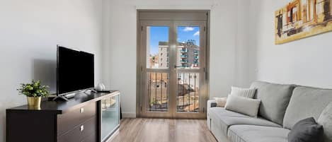 A modern living room with a flat-screen TV, a dark wooden dresser, a grey sofa, and a large window with a view of buildings.