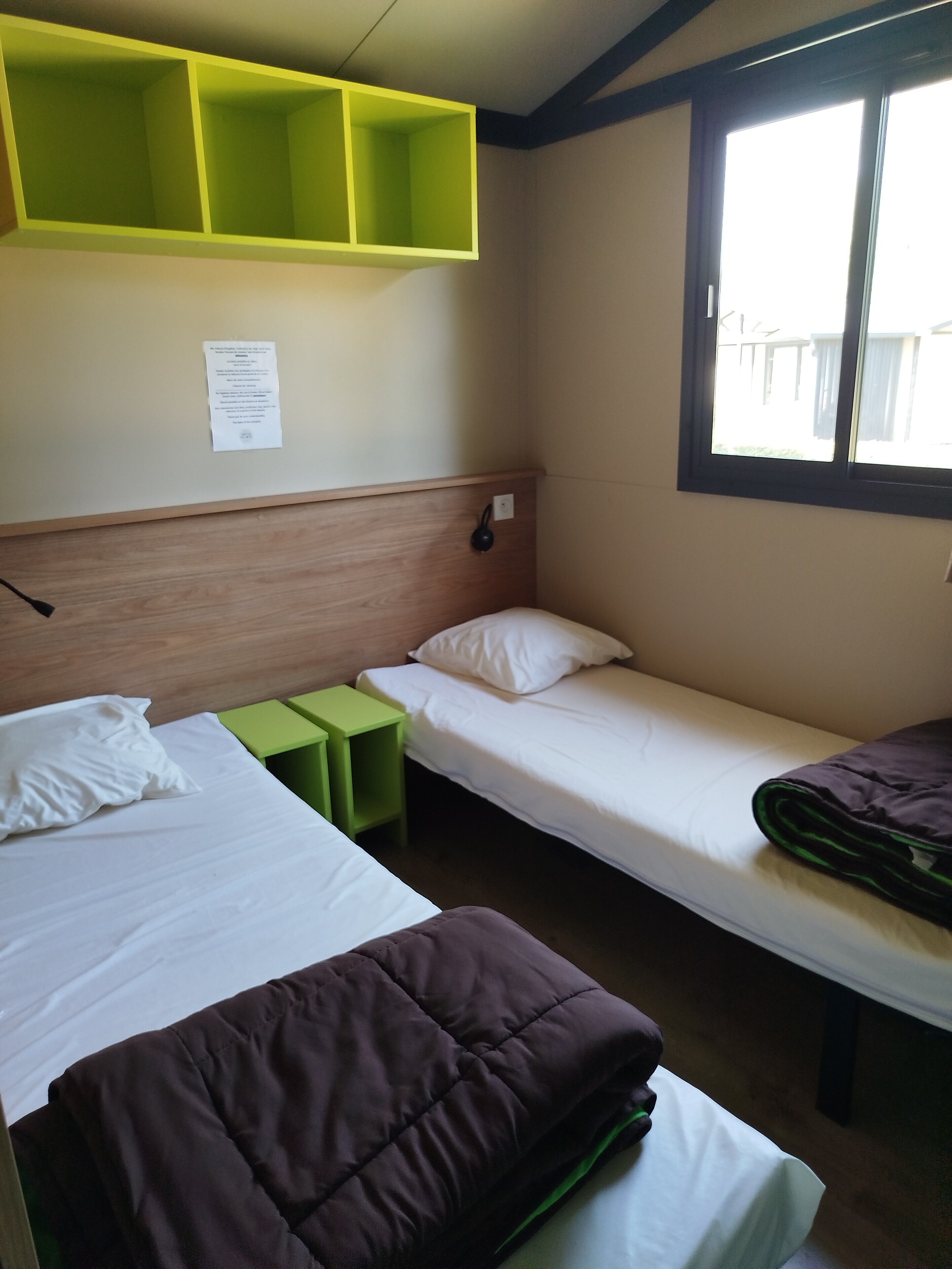 Ein Zimmer mit zwei Einzelbetten, einem grünen Nachttisch, einem Holz-Kopfteil, einem Fenster mit Blick und einer Wandleuchte.