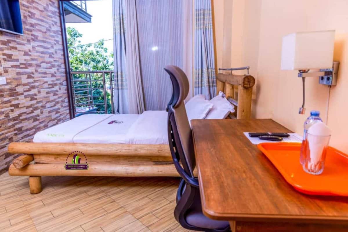 A room with a bed, a wooden table, a chair, a window with curtains, and a view of greenery outside.