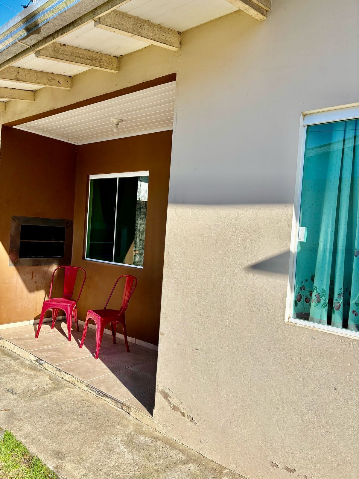 A building with a covered patio area, two red plastic chairs, a window with a curtain, and a television mounted on the wall.