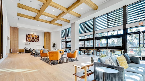 A spacious lobby with a wooden floor, a grey sofa, orange chairs, and large windows with blinds.