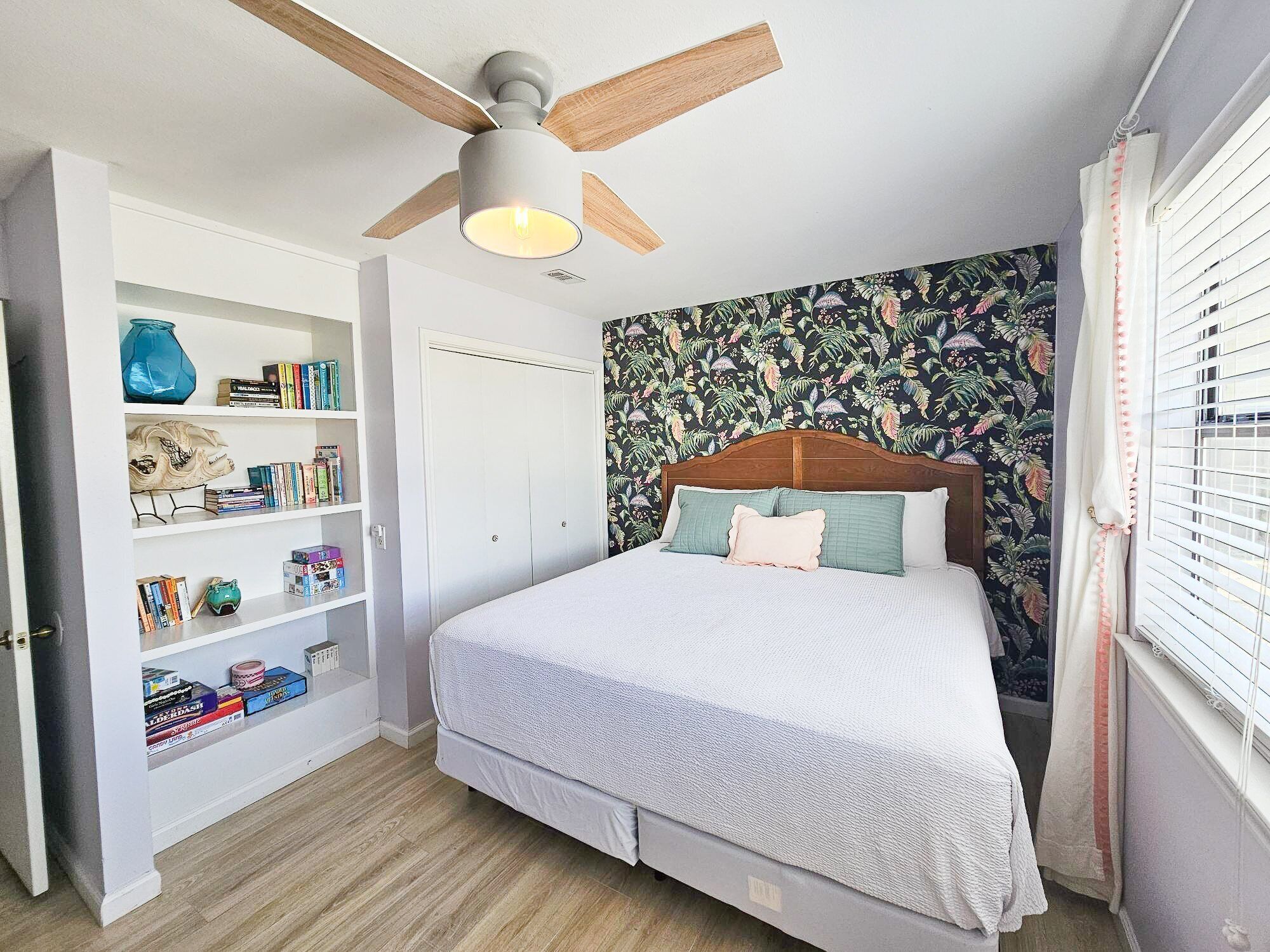 Une chambre à coucher avec un lit, un ventilateur de plafond, une bibliothèque et un papier peint à motifs floraux.