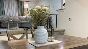 A dining area with a wooden table, cushioned benches, a white vase with green plants, and a grey sofa in the background. - Beautifully updated 3BR near Airport/RIT/UofR/mcc (Rochester)