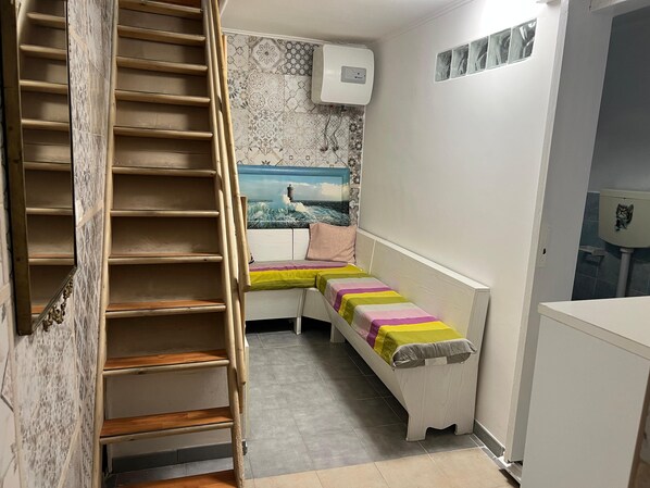 Una habitación estrecha con una escalera de madera, un banco con cojines de colores, un aire acondicionado de pared y un piso de baldosas.