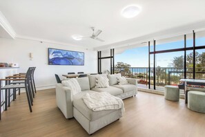 A spacious living area with a sofa set, a dining table with chairs, a ceiling fan, and a large window offering a view of the outdoors.