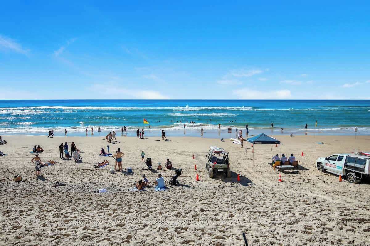 Une plage où des gens se prélassent au soleil et se baignent, un poste de surveillance des sauveteurs et un véhicule de sauvetage.