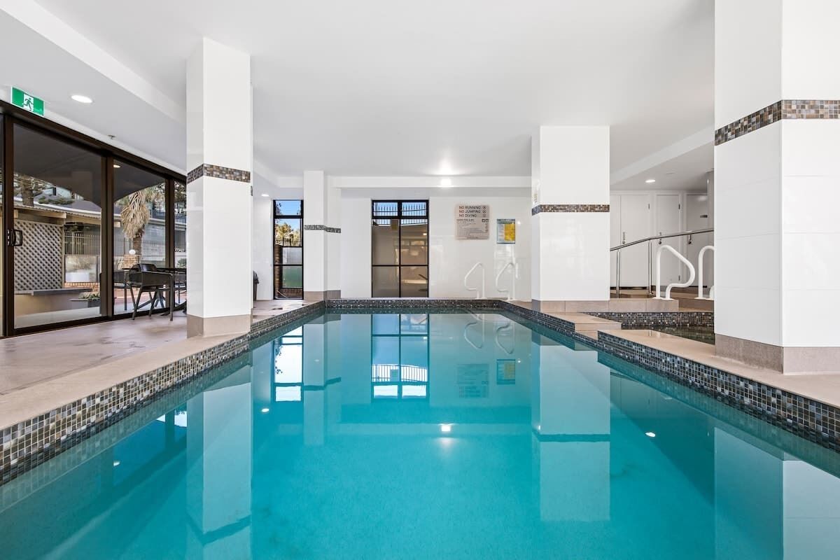 Une grande piscine intérieure à l’eau claire, entourée de murs et de colonnes blancs, et dotée d’une entrée avec une porte en verre.