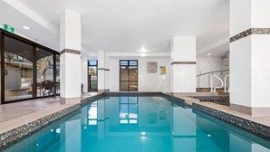 Un'ampia piscina interna con acqua cristallina, circondata da pareti e colonne bianche e un ingresso con porta in vetro.