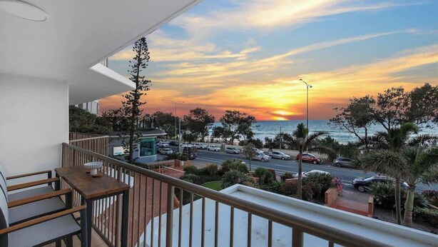 Un balcone con vista sul tramonto sul mare, una strada con auto parcheggiate e della vegetazione.