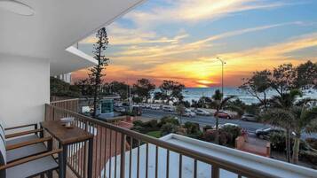 Ein Balkon mit Blick auf den Sonnenuntergang über dem Meer, eine Straße mit geparkten Autos und etwas Grün.
