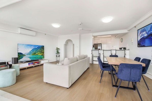 A modern living room with a flat-screen TV, a sofa, a dining area with chairs, and a kitchen in the background.