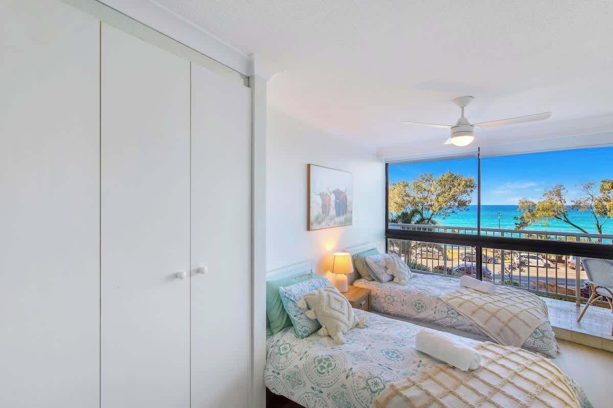 Une chambre avec deux lits, un ventilateur de plafond, une table de chevet avec une lampe et une grande fenêtre donnant sur une plage.