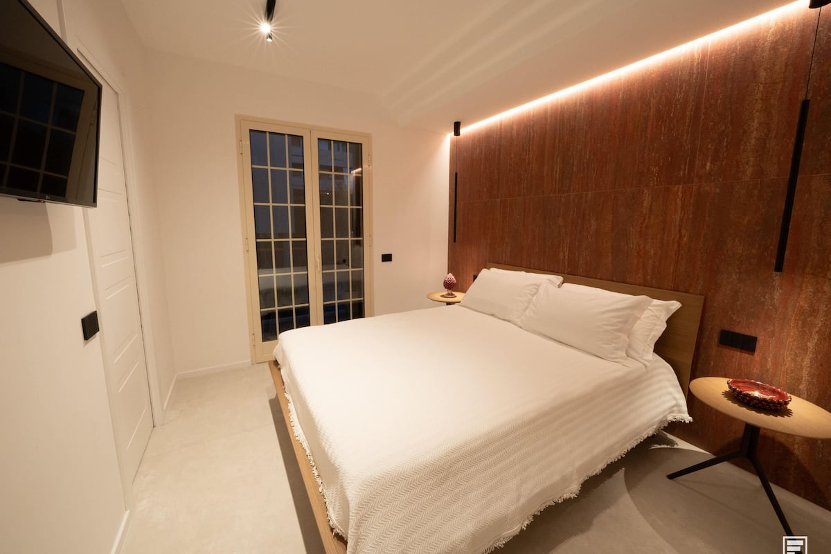 Une chambre à coucher moderne avec un grand lit, un téléviseur fixé au mur et une porte-fenêtre coulissante.
