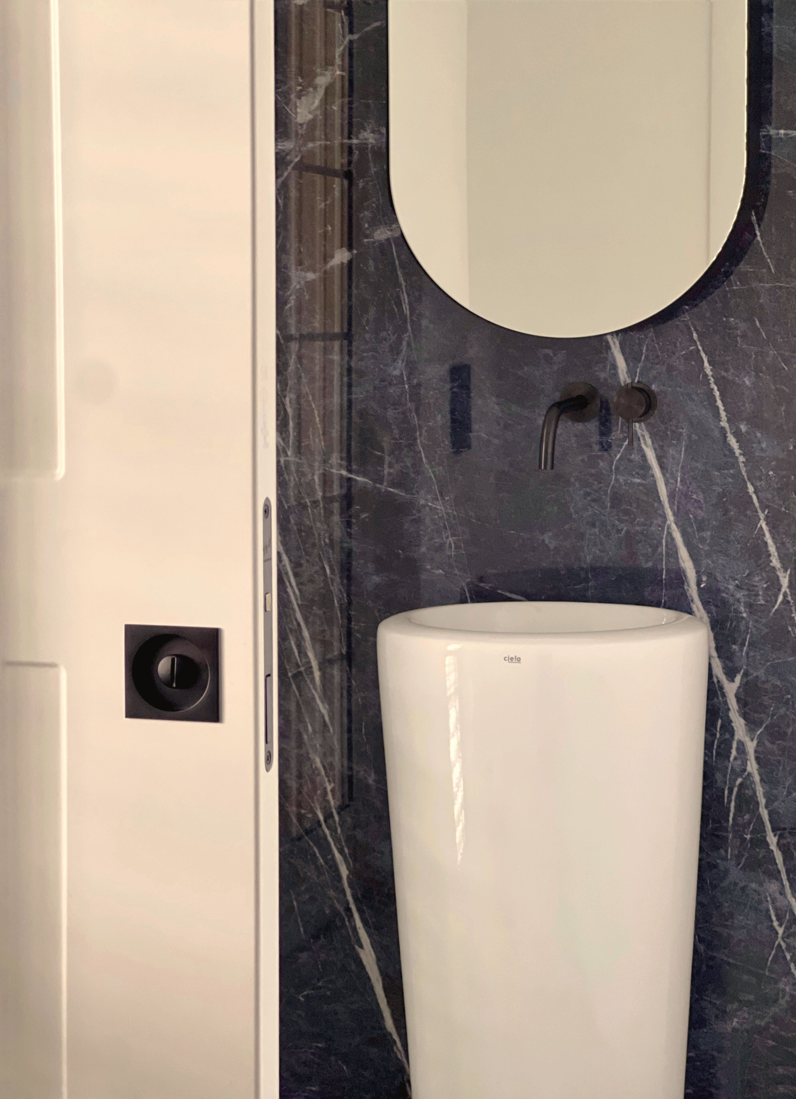 Un baño con puerta blanca, un lavamanos blanco y un espejo con marco negro.