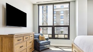 Un dormitorio moderno con un ventanal grande, un televisor de pantalla plana montado en la pared, un aparador de madera, un sillón de cuero y una cama con edredón blanco.