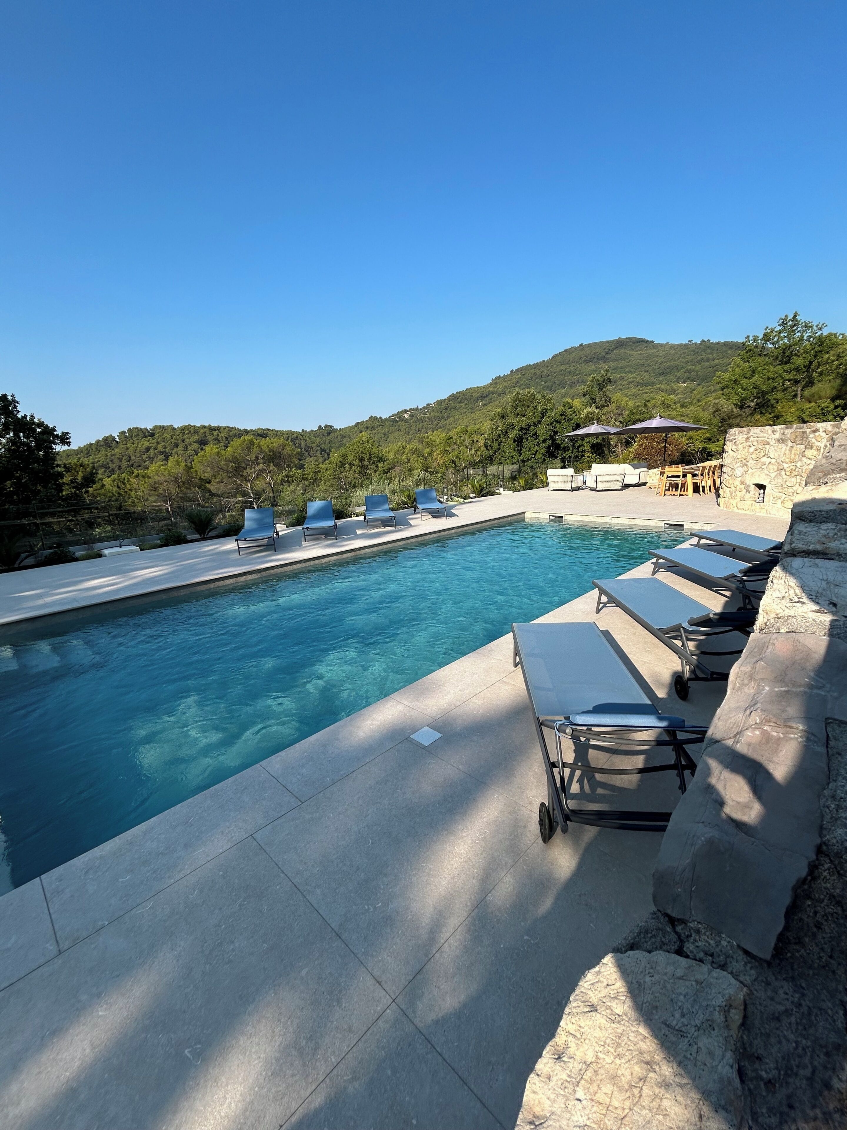 Zona de piscina amb gandules i ombrel·les, envoltada de murs de pedra i una vista a un turó forestal.