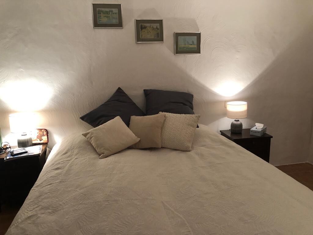 A neatly made bed with a beige comforter and several pillows, a bedside lamp, and a small table with a lamp and a picture frame.
