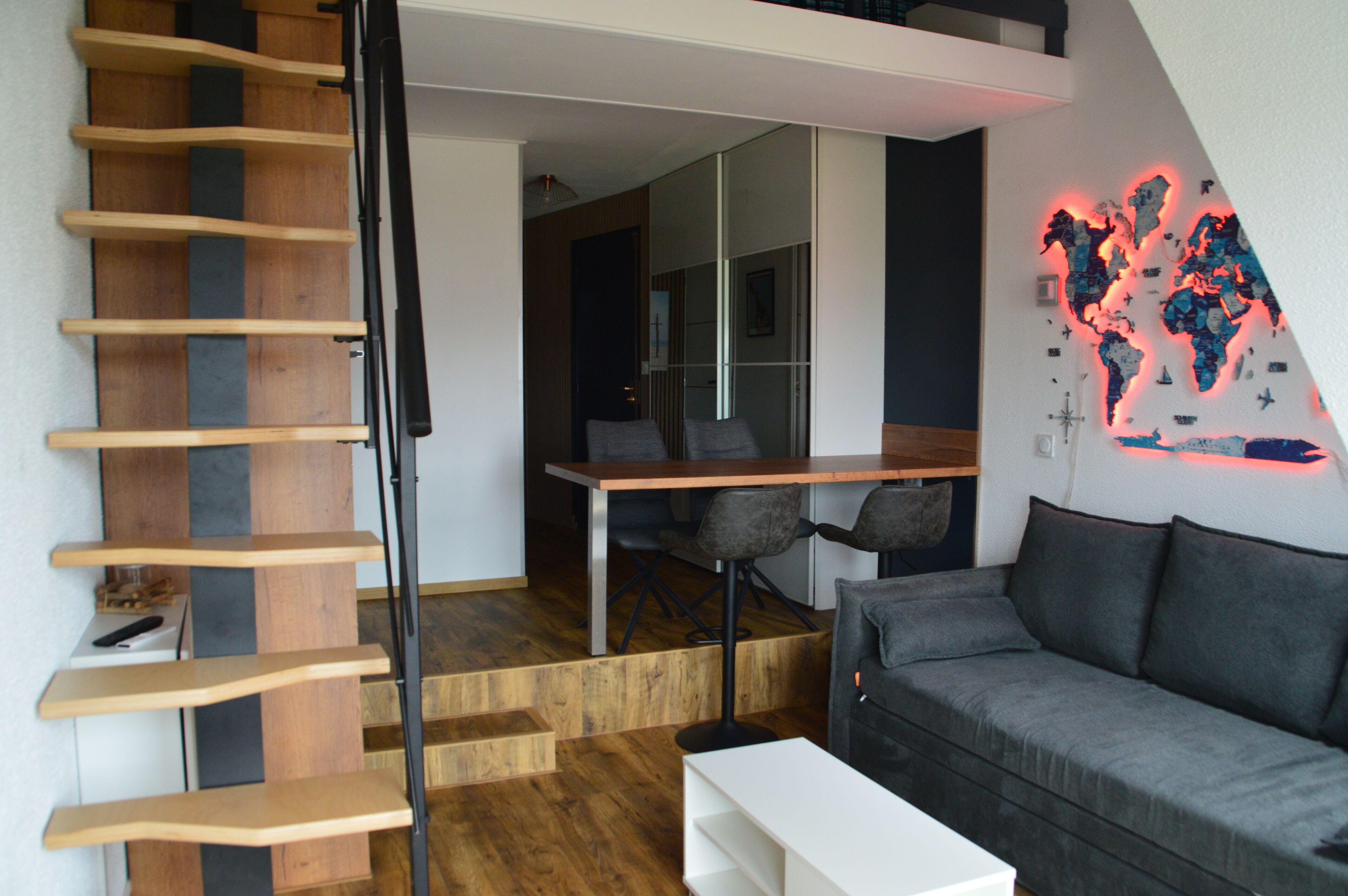 A modern interior with a wooden staircase, a grey sofa, a dining table, and a world map mural.
