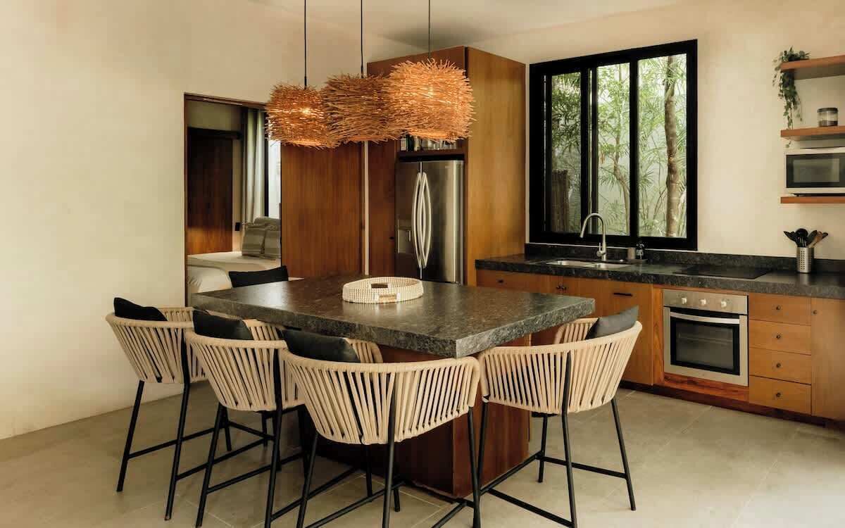 A modern kitchen with a central island, bar stools, a stainless steel refrigerator, a microwave, and a window with a view of trees.