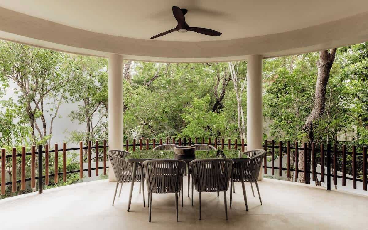 A covered outdoor seating area with a round table and chairs, surrounded by a wooden fence, overlooking a lush green forest.