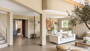 Ein geräumiger Wohnbereich mit einem weißen Sofa, einem Couchtisch und einer Treppe. An den Wänden befinden sich Kübelpflanzen und eine große Holzskulptur.