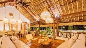 Un área de descanso interior con suelo de madera, asientos acolchados, lámparas de mimbre colgantes y vistas a plantas tropicales.