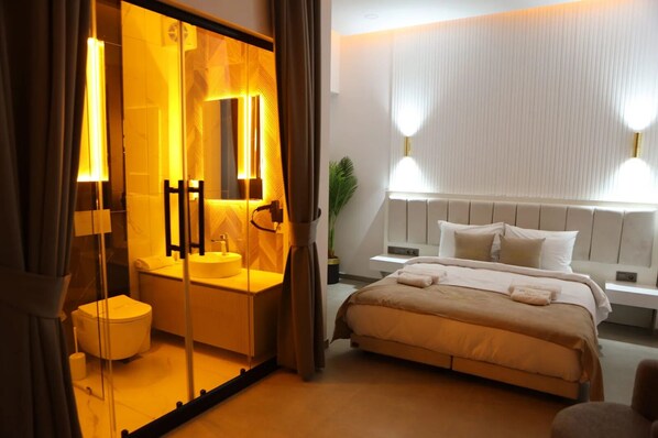 Une chambre d’hôtel avec un grand lit, une table de chevet, une commode, un miroir, un lavabo rond et des toilettes.