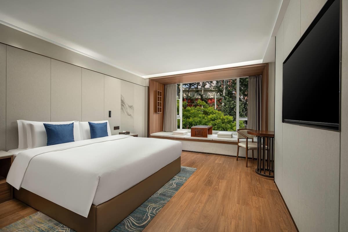 A modern hotel room with a large bed, a desk, and a window with a view of greenery.