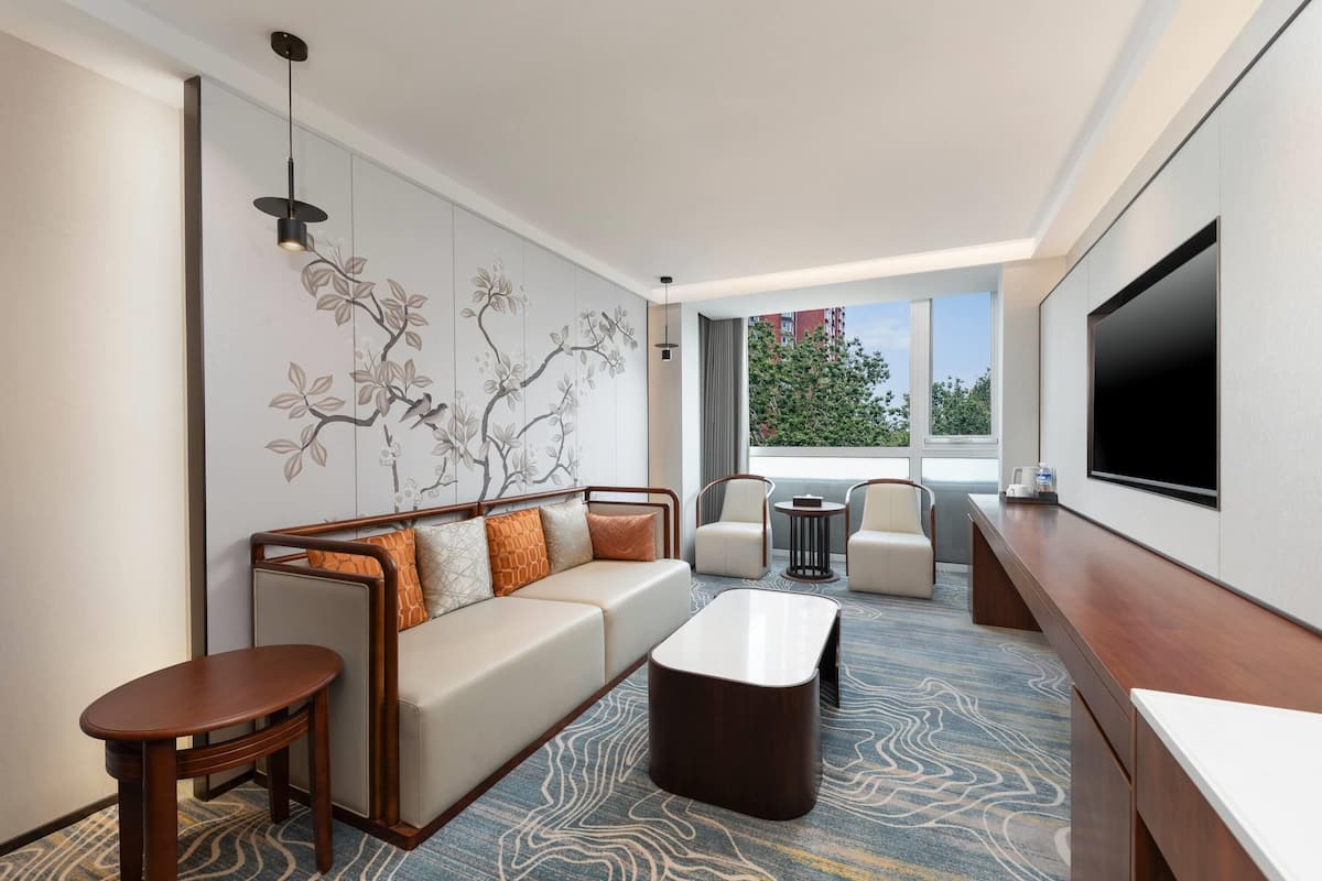 A modern hotel room with a sofa, armchairs, a coffee table, and a TV.