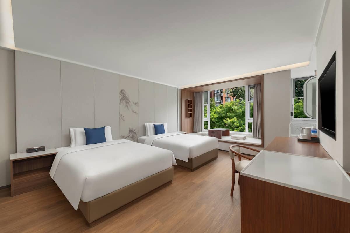 A hotel room with two beds, a desk, a TV, and a window with a view of greenery.