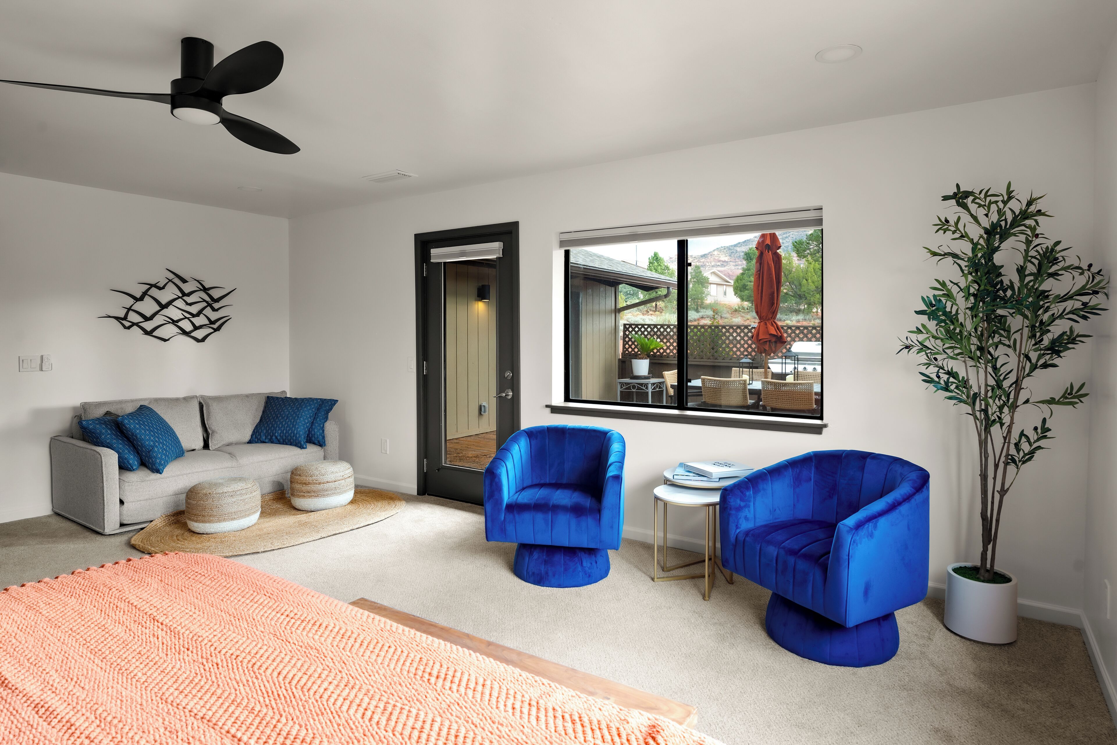 Een moderne woonkamer met een bank, twee blauwe fauteuils, een salontafel, een groot raam en een plafondventilator.
