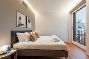 A modern bedroom with a large bed, a bedside table with a water bottle, and a view of the outside balcony.