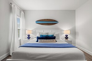A bedroom with a bed, two bedside tables, a wall-mounted decorative piece, and a window with curtains. - Beach House - Redfish Cove by PHG (Port St. Joe)